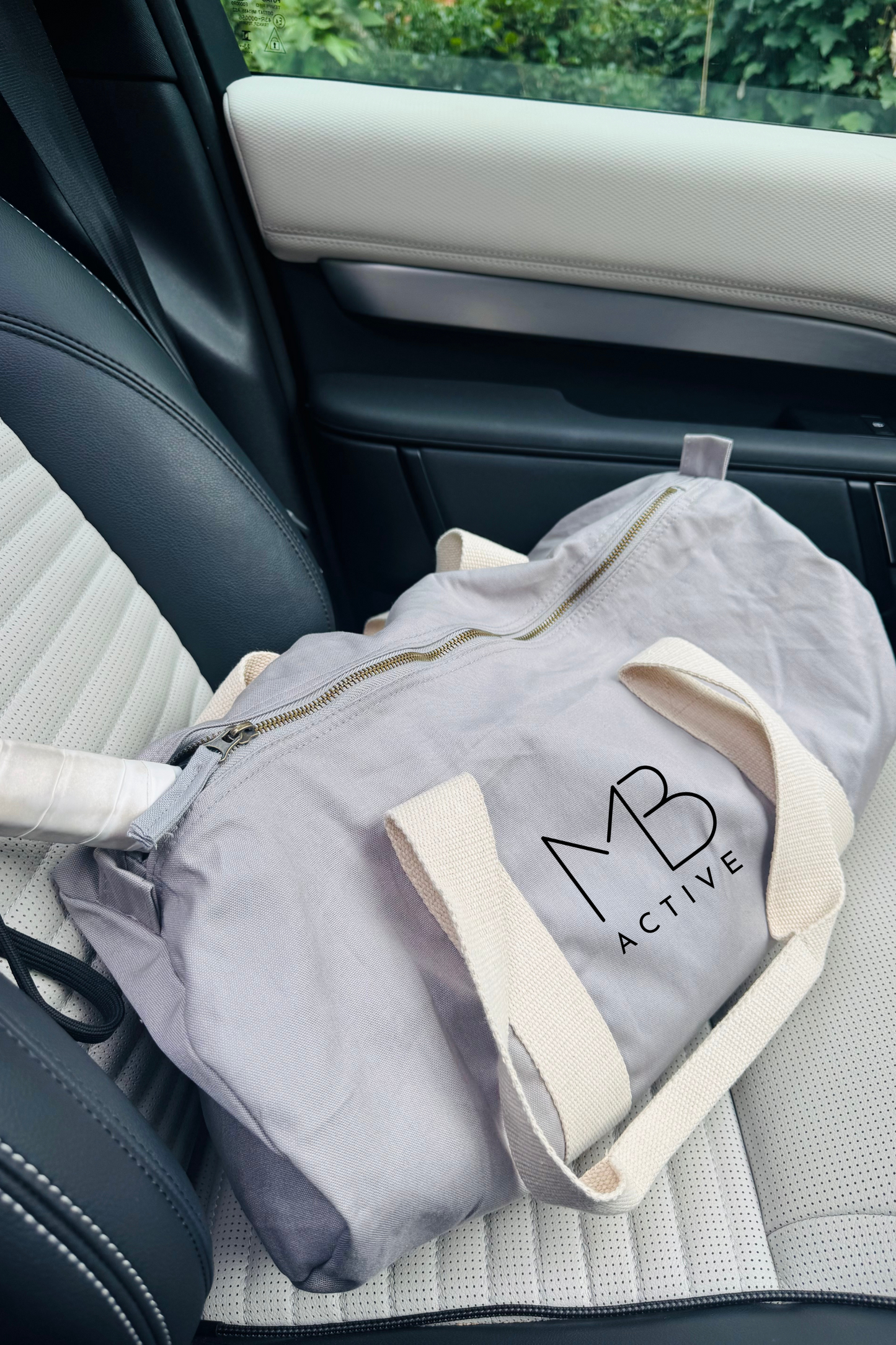 Light grey organic cotton SOL barrel bag with beige handle on a car seat, featuring 'MB Active' logo branding by Meraki Beach.