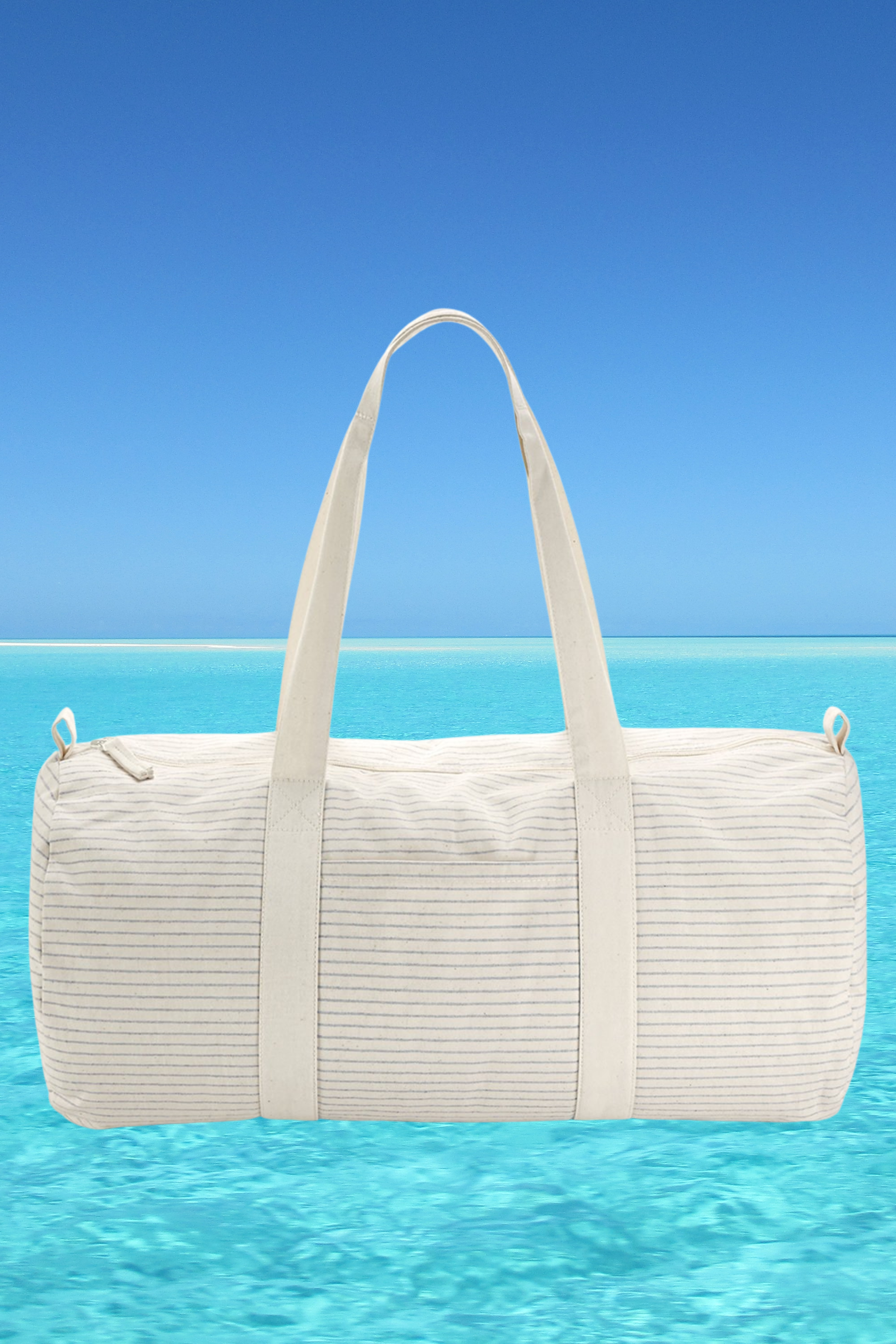 The Meraki Beach CORA organic cotton striped barrel bag in light grey with beach back drop