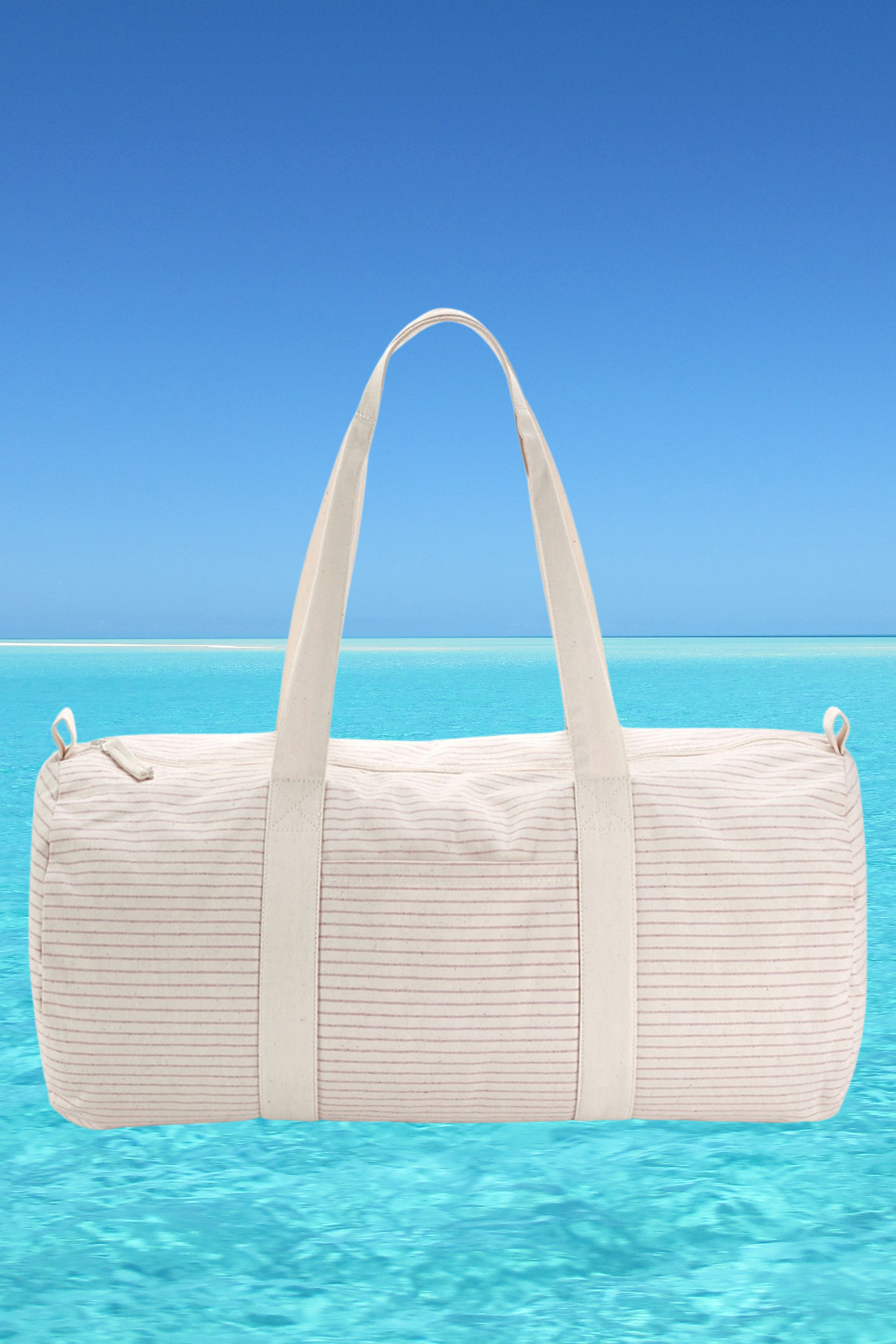 The Meraki Beach CORA organic cotton striped barrel bag in light red with beach back drop