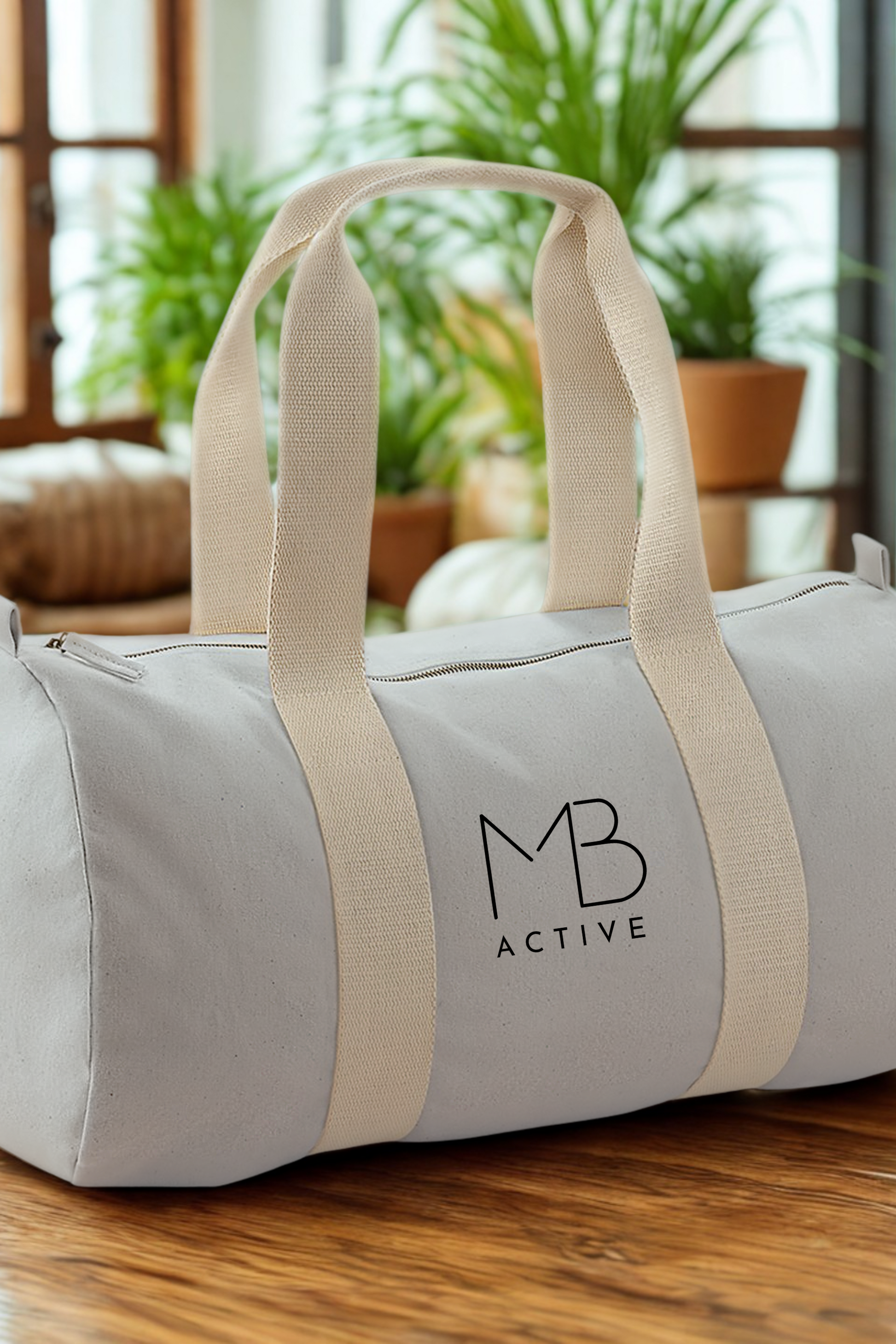 Light grey SOL organic cotton barrel bag with cream straps and 'MB Active' logo on a wooden surface with plants in the background by Meraki Beach. 