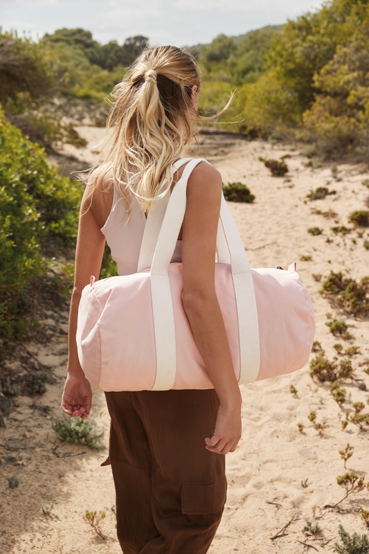 Lady walking on beach with a Meraki Beach organic cotton SOL barrel bag in pastel pink with cream straps. 